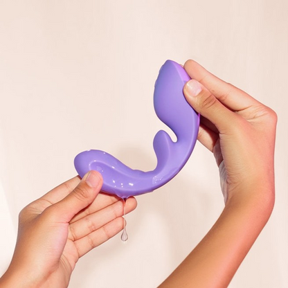 Hands holding a purple Flow, Bedroom Toy Vibrator by Liberator with water on it against a white background.